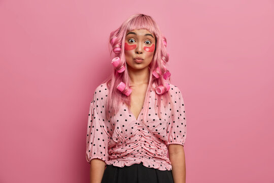Photo of surprised long haired woman wears hair curlers, makes perfect hairstyle, looks with shocked expression, applies collagen pads under eyes, dressed in polka dot blouse, gets ready for going out