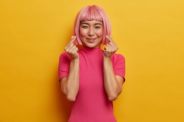 Gentle smiling pretty woman with trendy pink haircut, makes korean like gesture, expresses love, being in good mood, dressed in bright rosy t shirt, isolated on yellow wall. Body language concept.