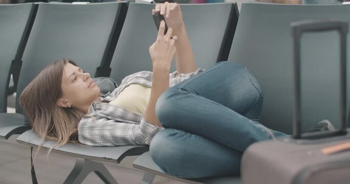 Bored tired young woman lying on chairs in airport waiting area and using smartphone. Portrait of brunette beautiful Caucasian tourist wait for delayed departure. Cinema 4k ProRes HQ.