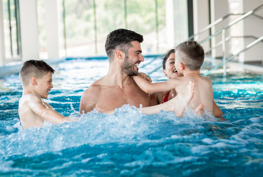 Smiling Family Of Four Having Fun And Relaxing In Indoor Swimming Pool At Hotel Resort.