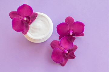 White container with cream for face and body with three magenta colored orchid flowers on purple background. Concept of delicate facial cosmetics with orchid extract
