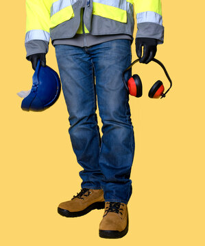 Workman  Wearing Jeans, Gloves, Boots And Holding Protective Equipment, Helmet And Ear Defender   On Yellow Background