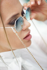 Fototapeta premium Close-up Portrait of a young beautiful fashionable woman in sunglasses. A lady poses in the open air. The model is dressed in a stylish white jacket. Women's fashion.Toned