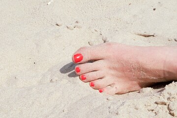 Female left foot on the sand, on the beach. Nails painted with red varnish.