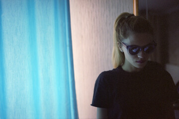 Close-up of a young caucasian CIA woman in black sun goggles standing in a room near blue curtains. Istanbul, Turkey.  Scanned film - obrazy, fototapety, plakaty
