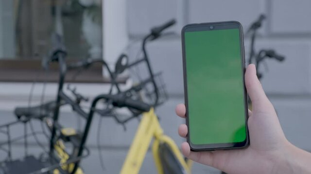 Female hands holding black smartphone with green screen on city street BG.