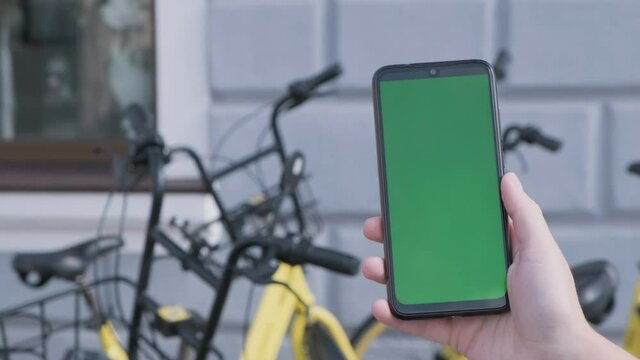 Female hands holding black smartphone with green screen on city street BG.