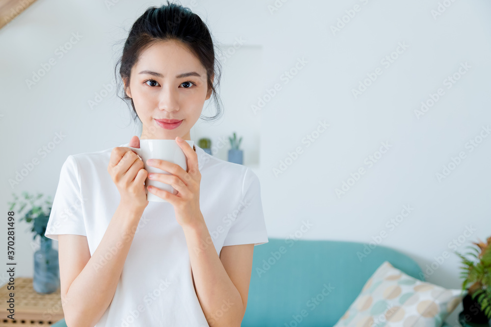 Wall mural Young asia woman with ponytail in white t-shirt holding a cup of coffee or tea relaxing at home in the morning.