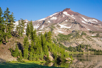 Volcanic mountain landscape with clear blue sky, morning light, and reflective water in the foreground.