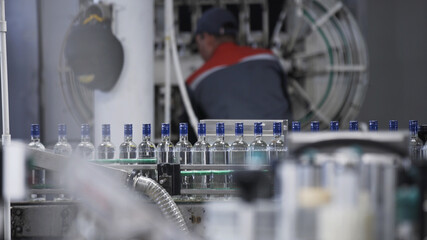 Transparent glass bottles sealed with blue foil and filled with alcohol move along conveyor line with control panel in Factory for bottling vodka. Engineers set up bottling machine in background.