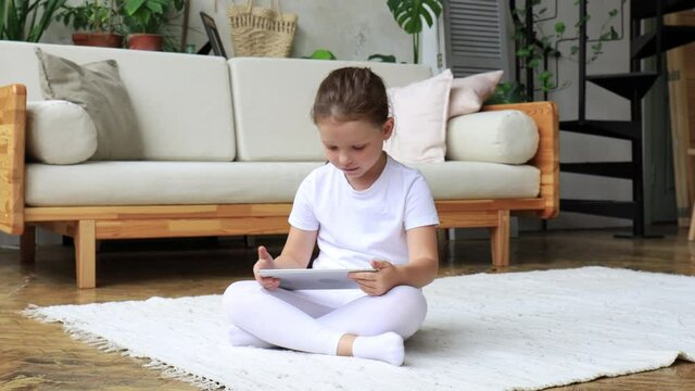 Child girl using digital tablet lying on carpet floor alone, holding and surfing touchpad internet at home.