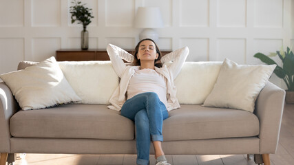 Fototapeta premium Front view relaxed young 30s woman sleeping on comfortable couch with folded hands behind head, enjoying weekend leisure lazy time, napping daydreaming or meditating alone in modern living room.