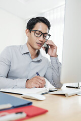 Young Asian entrepreneur talking on phone with business partner and writing in planner