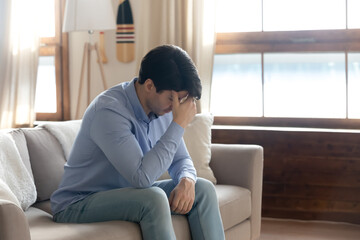 Side view stressed young guy touching forehead, feeling unwell alone in living room. Depressed millennial handsome man thinking of life problems, worrying about betrayal relations breakup at home.