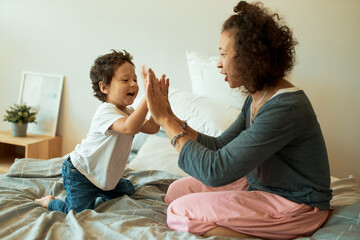 Happy mother and baby boy playing games at home. Attractive cheerful Latin female clasping hands with her infant son sitting on bed, enjoying nice time together. Fun, leisure and activity concept