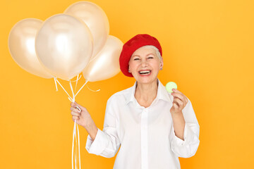 Colorful isolated image of overjoyed ecstatic middle aged European woman wearing white blouse and red beret laughing, having fun, holding helium balloons, eating macaron, being in good mood