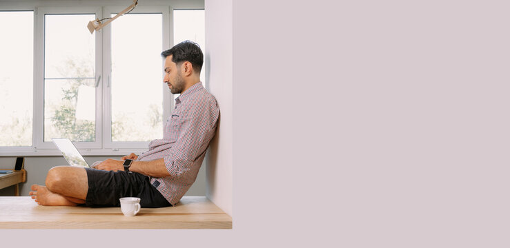 A young man in a shirt sits on the windowsill and works on a laptop at home
