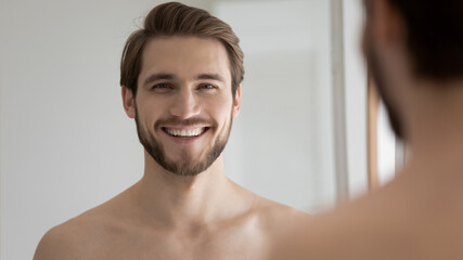 Head shot portrait close up mirror reflection smiling bearded young man with bare shoulders looking at camera, standing in bathroom, satisfied positive handsome guy enjoying morning routine