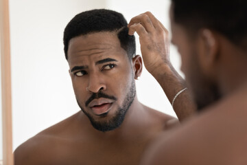 Close up anxious unhappy African American man checking hair after shower, looking in mirror,...