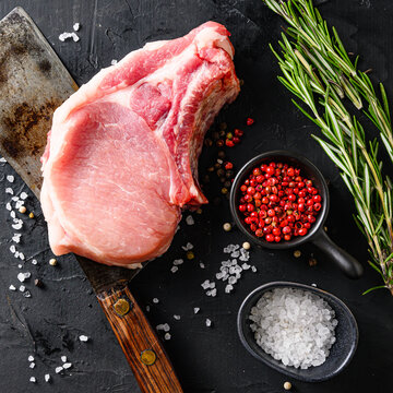 Organic cutlet on a rib or Pork meat over american classic butcher knife or cleaver with spices and rosemary and red pepper on black slate table top view. Square.