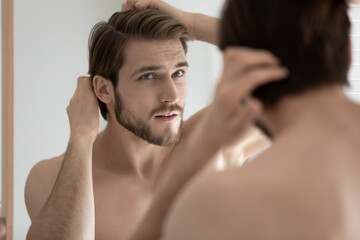 Close up head shot anxious stressed young man touching head, checking hair, looking in mirror, standing in bathroom, worried about dandruff or hair loss, healthcare and beauty concept