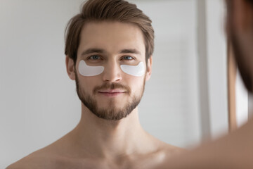 Head shot portrait mirror reflection close up handsome young man with hydrogel eye patches standing in bathroom at home, satisfied bearded guy with perfect skin enjoying skincare procedure