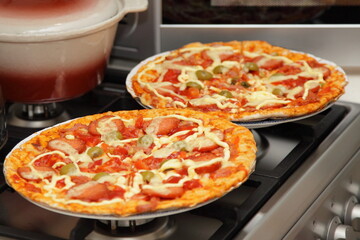 Two homemade pizza close up with sausages, cheese, green olives, ketchup and mayonnaise on gray Gas stove against the background of a microwave oven and pot, DIY kitchen