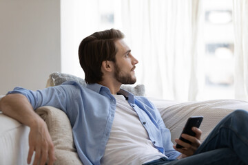 Dreamy thoughtful young man relaxing, holding smartphone, enjoying leisure time at home, peaceful...