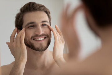 Close up mirror reflection smiling satisfied young man touching skin under eyes, applying anti...