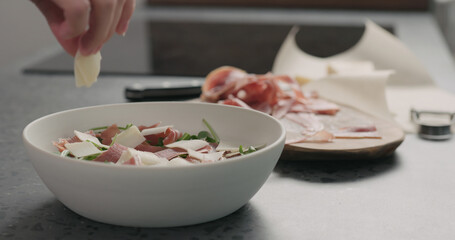 man hand adding parmesan flakes to make salad with arugula, tomatoes and prosciutto