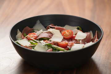 Salad with prosciutto, tomato, arugula leaves and parmesan cheese in black bowl on walnut