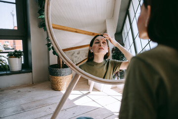 Attractive young caucasian woman in headphones touching face and looking at mirror in living room at home in sunny day. Beauty and skin care concept.