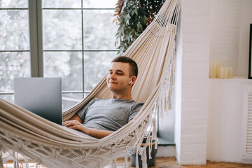A young caucasian businessman in headphones works on a laptop, browses the internet while lying in a hammock at the home office. A person working on electronic devices.