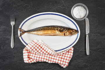 Whole smoked mackerel fish on white oval plate, gray black stone like table under. View from above
