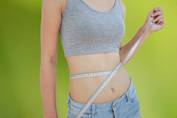 A slender tanned girl on a green defocused background, holding a centimeter with her hands around...