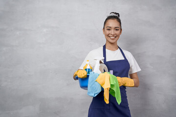 Young happy woman, female cleaner in uniform and rubber gloves holding bucket of cleaning supplies...