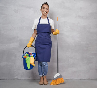 Happy cleaning lady in uniform and yellow rubber gloves holding broom and plastic bucket with rags and different cleaning products while standing against grey wall