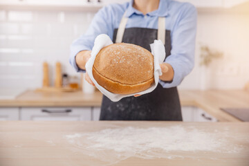 Hands baker holding round loaf of white bread. Woman in apron in home Scandi kitchen background