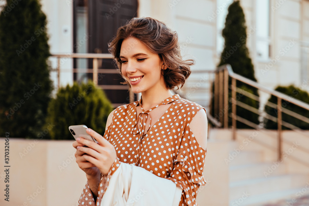 Wall mural Stunning female model in elegant blouse looking at phone screen with interested face expression. Outdoor photo of pleased european woman in brown attire texting message with smile.