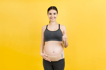 Portrait of pregnant woman wearing maternity belt on her belly and showing thumb up gesture at yellow background. Orthopedic abdominal support belt concept. Copy space
