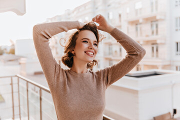 Stunning caucasian woman in cozy sweater laughing at balcony. Happy white girl with wavy hair enjoying life.