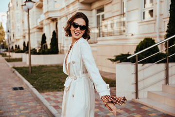 Good-humoured girl in long coat smiling on the street. Well-dressed european woman spending time in city.