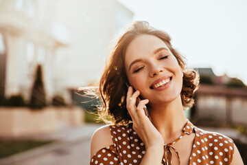 Glamorous young woman talking on phone with eyes closed. Outdoor photo of pretty caucasian girl...