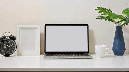 Fototapeta premium Mock up stylish of workspace with blank screen laptop, clock, frame, coffee cup and vase on white table and white background. Front view.