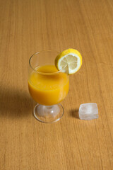 An elegant glass with a lemon wedge on the rim full of refreshing orange juice on a table with an ice cube on the side.