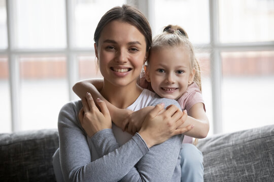 Cute little girl cuddling smiling beautiful young mother, sitting together on sofa, looking at camera. Portrait of affectionate millennial woman posing for photo with adorable preschool daughter.