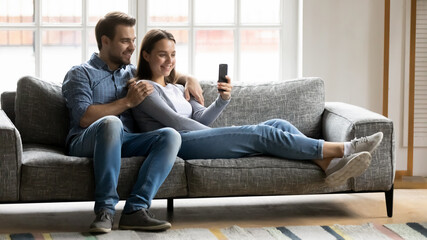 Obraz premium Full length relaxed young couple spending leisure time on comfortable sofa, watching funny videos in social network on smartphone, making video call or posing for selfie photo together indoors.