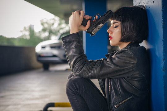 Beautiful brunette sexy spy agent (killer or police) woman in leather jacket and jeans with a gun in her hand running after someone, to catch him on parking