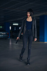 Beautiful brunette sexy spy agent (killer or police) woman in leather jacket and jeans with a gun in her hand running after someone, to catch him on parking