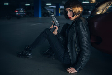Fototapeta na wymiar Beautiful brunette sexy spy agent (killer or police) woman in leather jacket and jeans with a gun in her hand running after someone, to catch him on parking